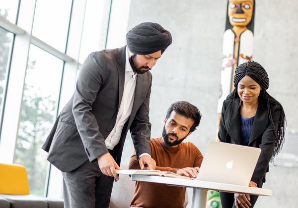 A group of 3 diverse, mature students are at a table with a laptop, working in s room with large, bright windows an a traditional totem in the background