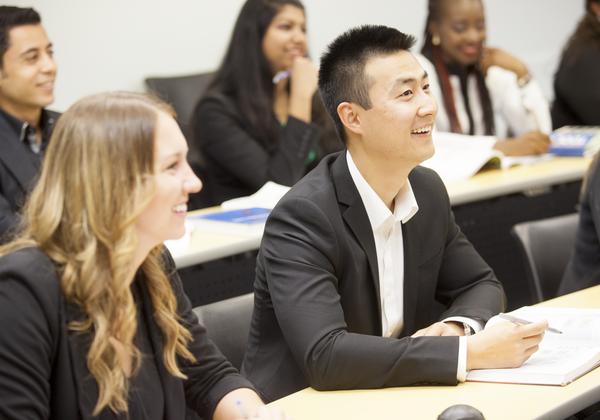 Five smiling students from divers backgrounds, dressed professionally, sitting at long rows in a classroom.