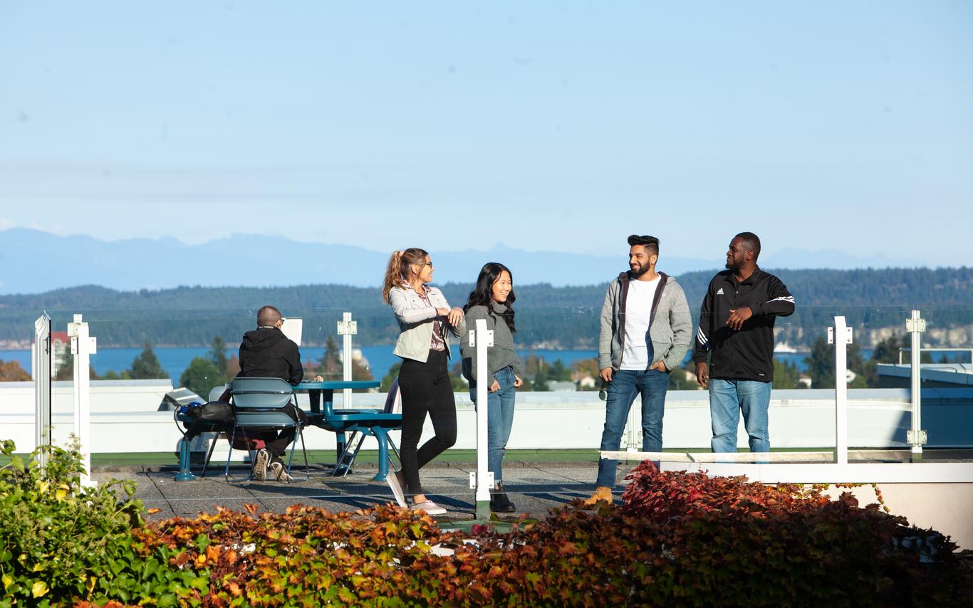 four students leaning on a railing on a rooftop patio with climbing leaves in the foreground and a person at a table and a mountain in the background