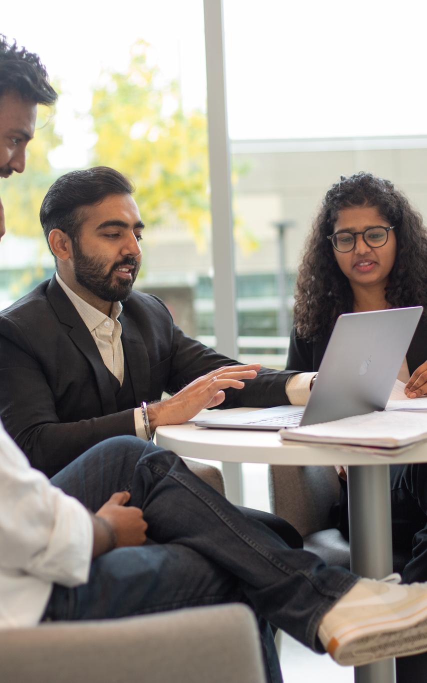 Four students in professional clothes are looking at a laptop sitting around a table.