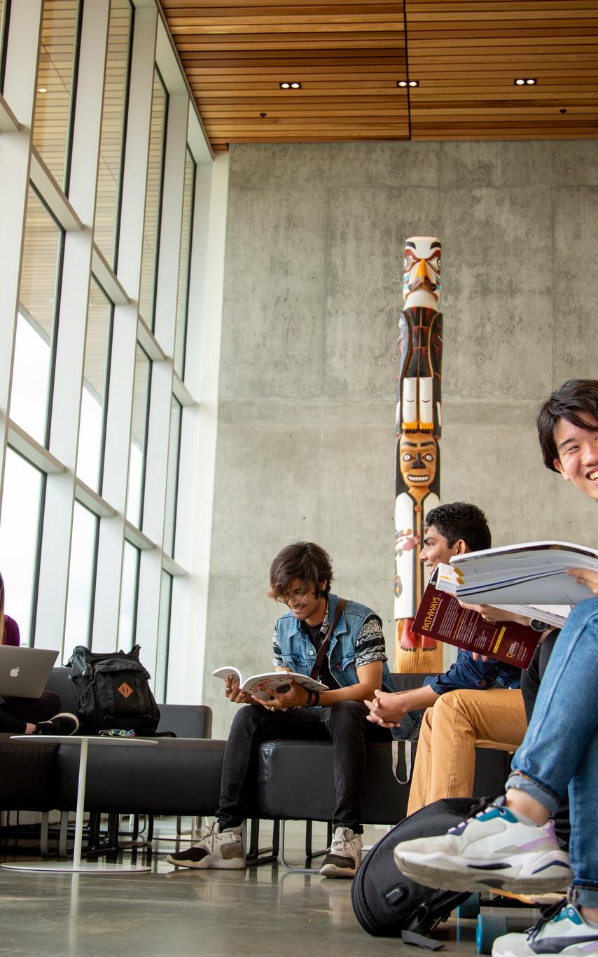 Students on lounge chairs in an open room with tall windows and a traditional totem in the background.